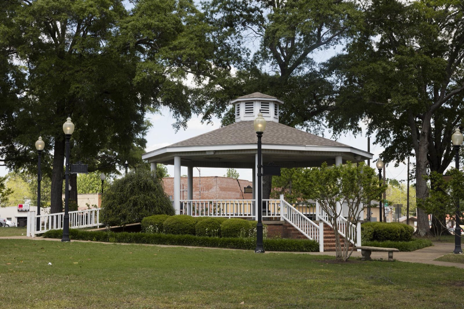 A white octagonal gazebo with lampposts and trimmed hedges stands under large shade trees on the town square in Calhoun City, Mississippi.