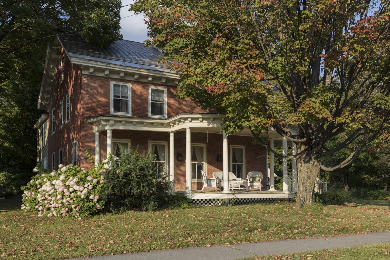 A red brick house with a white-columned wraparound porch, wicker chairs and pink hydrangeas sits under a maple tree in Chester, Vermont.