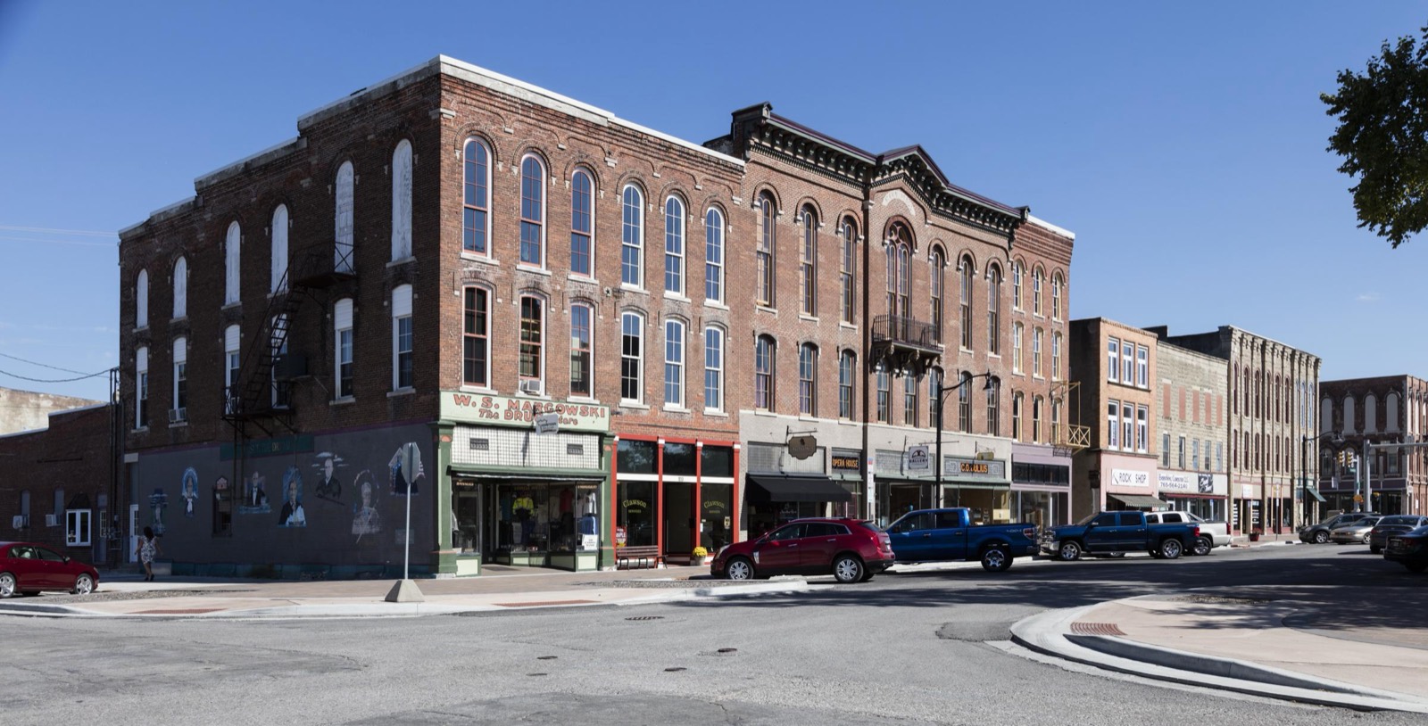 A block of three-story Victorian brick buildings faces the courthouse square in Delphi, Indiana, with cars parked along the curb.
