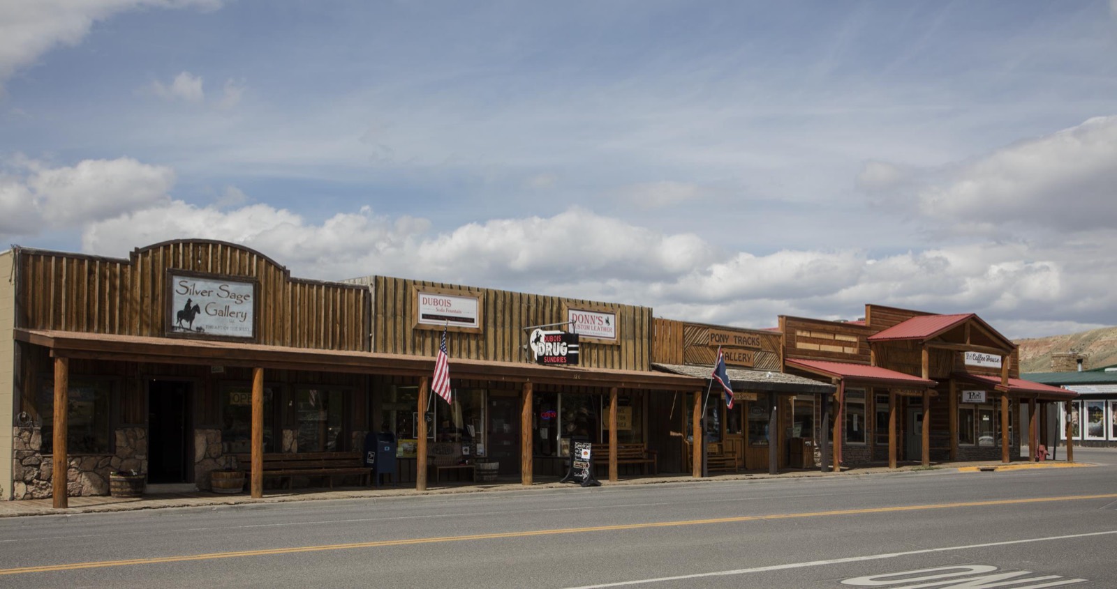 Rustic wooden storefronts with a covered boardwalk and flags line the main street of Dubois, Wyoming, under a partly cloudy sky.