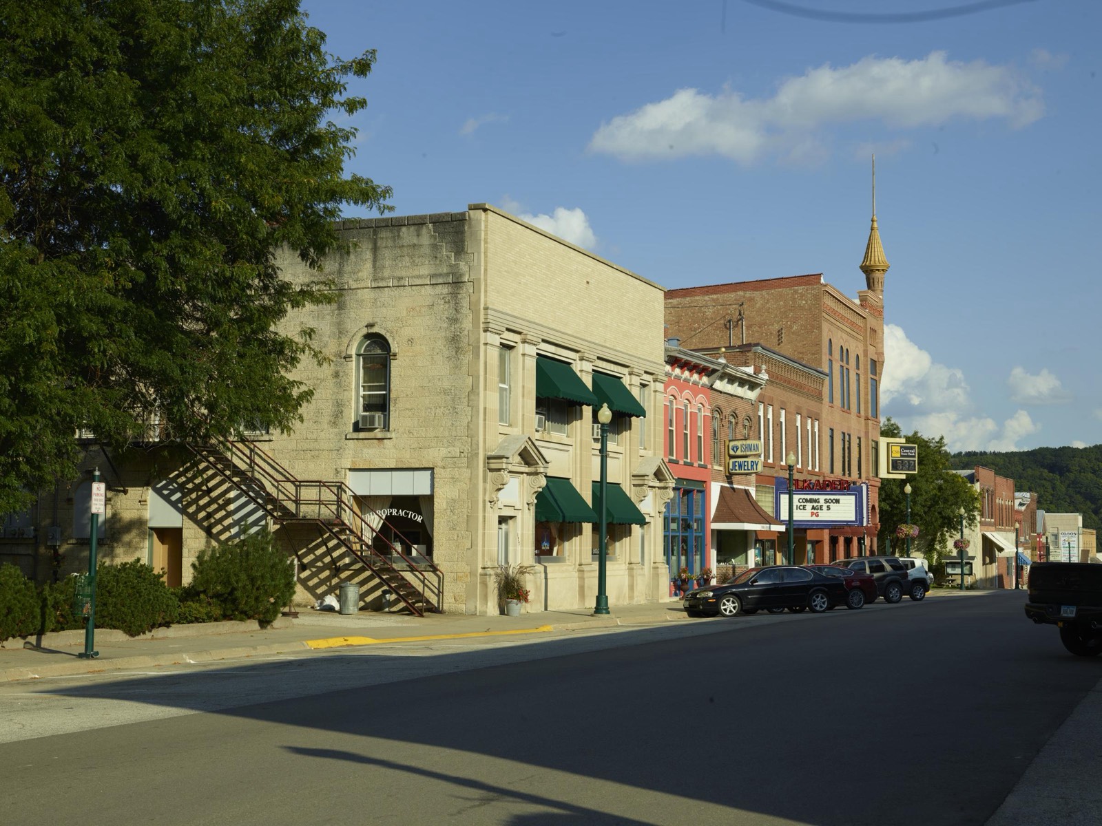 A row of historic brick and stone storefronts with green awnings and a small movie theater lines a sunny downtown street in Elkader, Iowa.