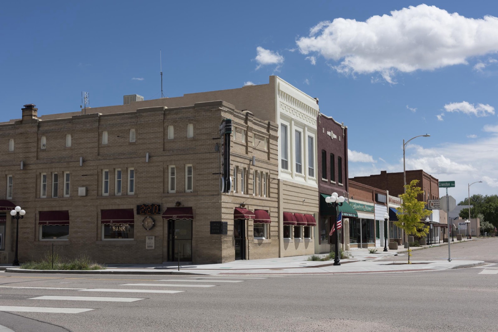 A tan brick bank building anchors a corner of Main Street in Fowler, Colorado, with a row of small shops beyond under a blue sky.