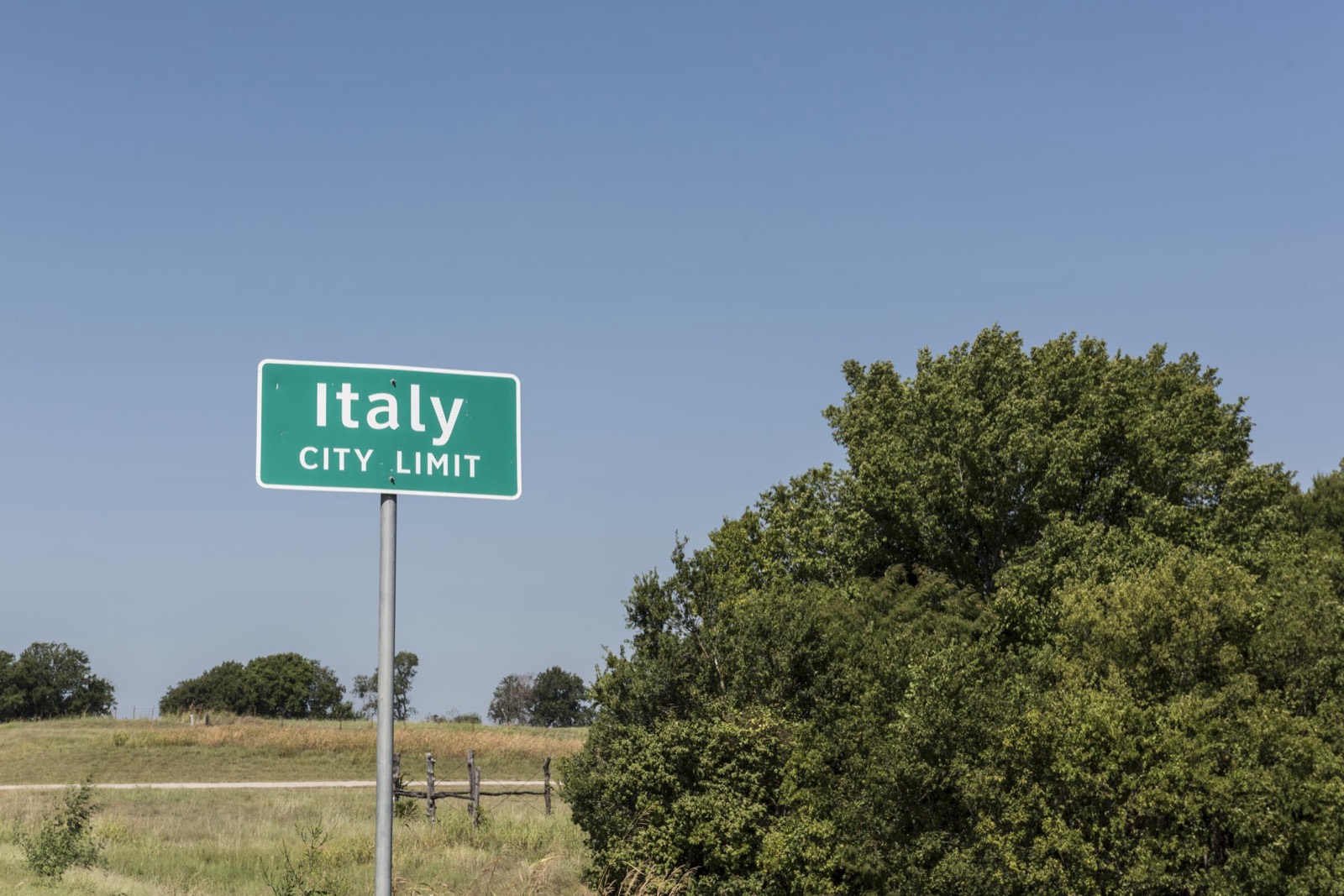 A green Italy City Limit sign stands beside pasture and leafy trees under a clear sky at the edge of Italy, Texas.