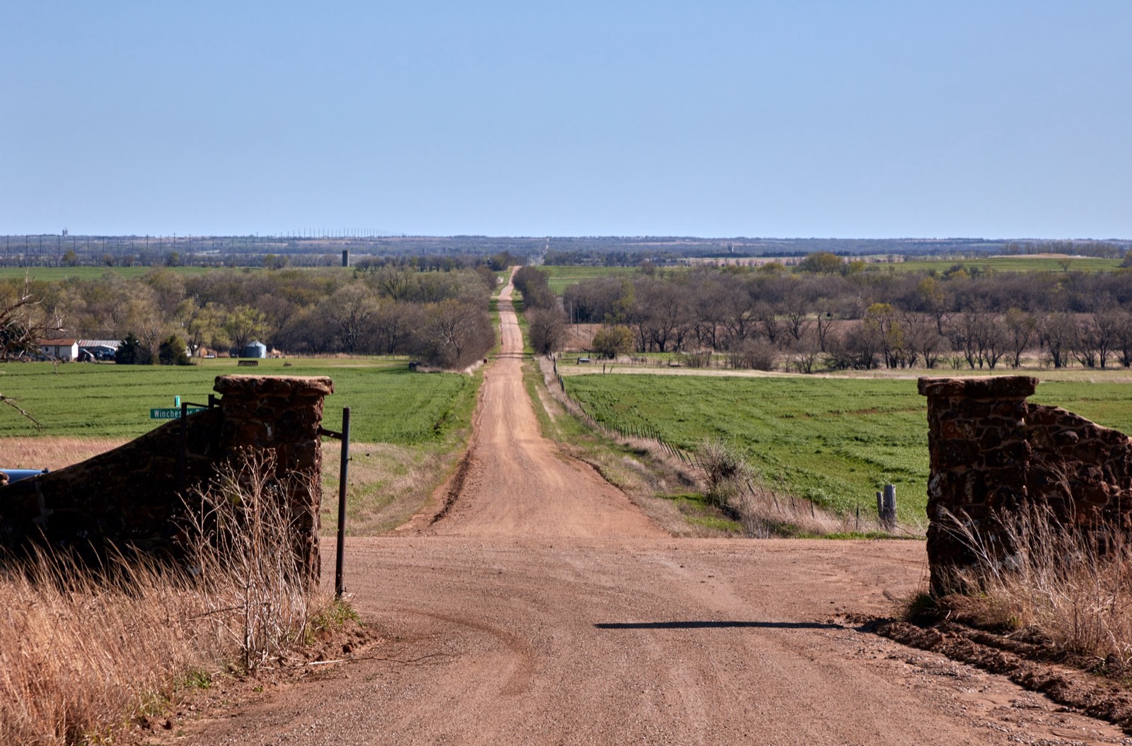 A long red dirt road runs straight between stone gateposts across green farmland near Lindsborg, Kansas.