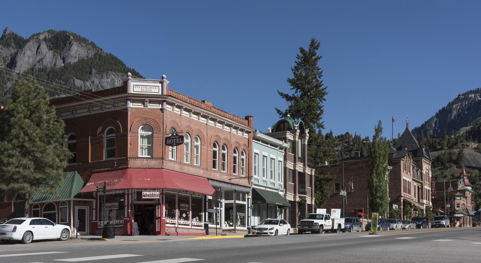 Brick storefronts and the historic Ouray Hotel line Main Street in Ouray, Colorado, beneath steep forested mountains and a clear blue sky.