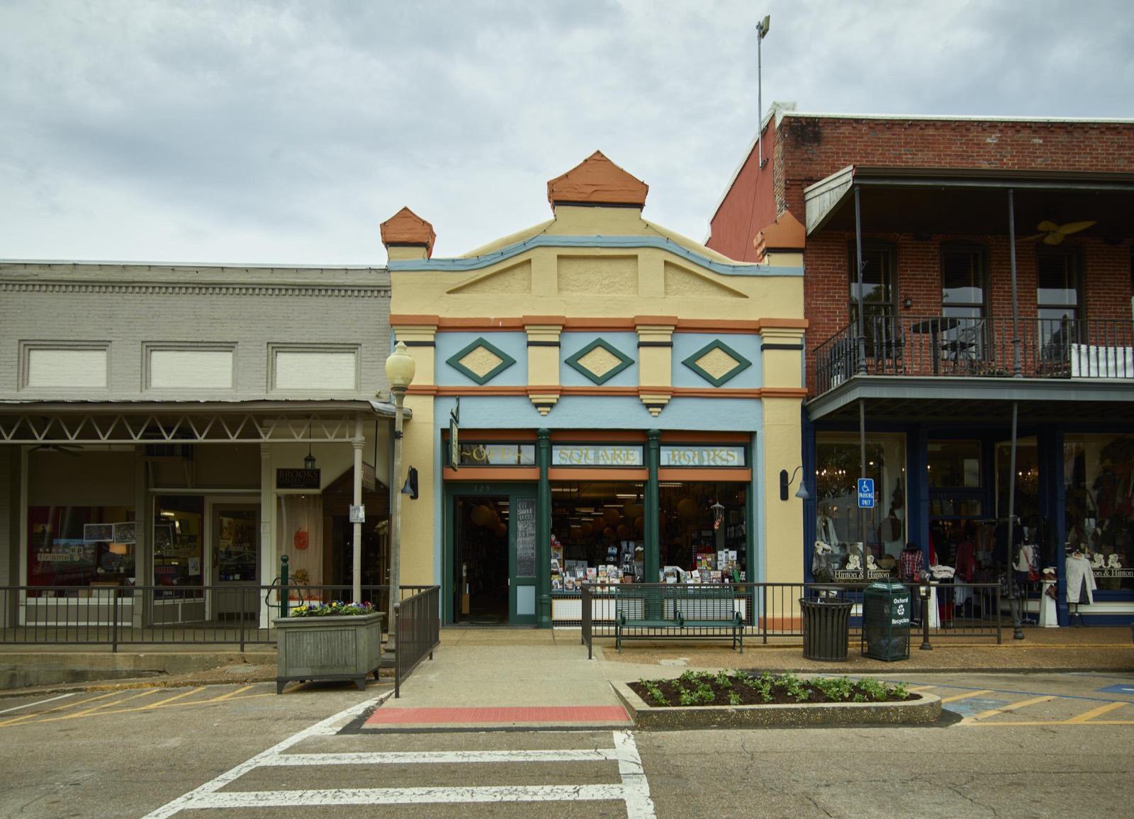 A colorful bookstore storefront sits between a gray shop and a brick building with an iron balcony on the town square in Oxford, Mississippi.