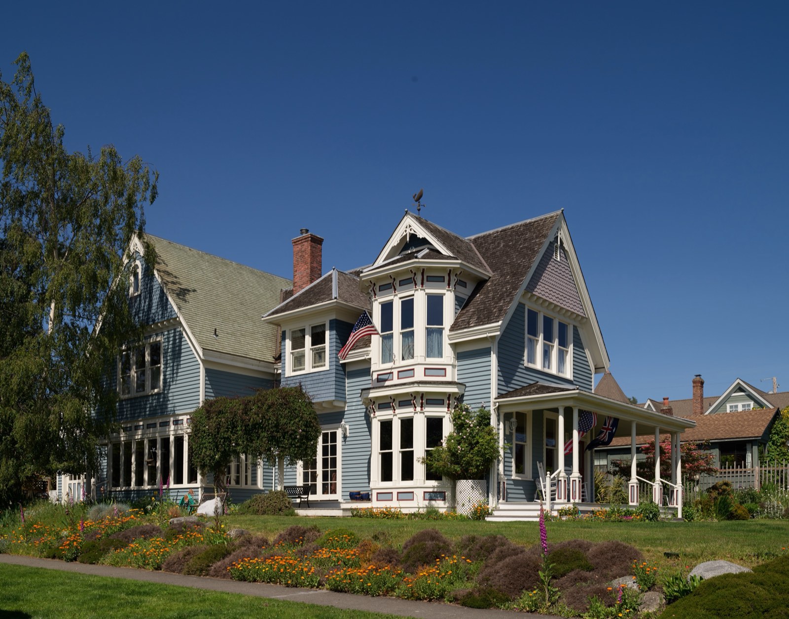 A blue Victorian house with white trim, bay windows and a wraparound porch rises above a flower-filled yard in Port Townsend, Washington.
