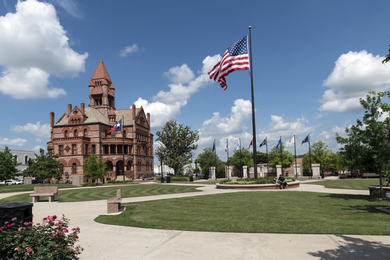 The red stone Hopkins County Courthouse stands beside a landscaped square with a large American flag in Sulphur Springs, Texas.