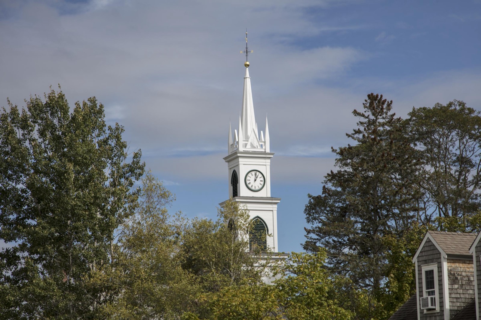 The white steeple and clock tower of the Tamworth Congregational Church rise above green trees against a blue sky in Tamworth, New Hampshire.