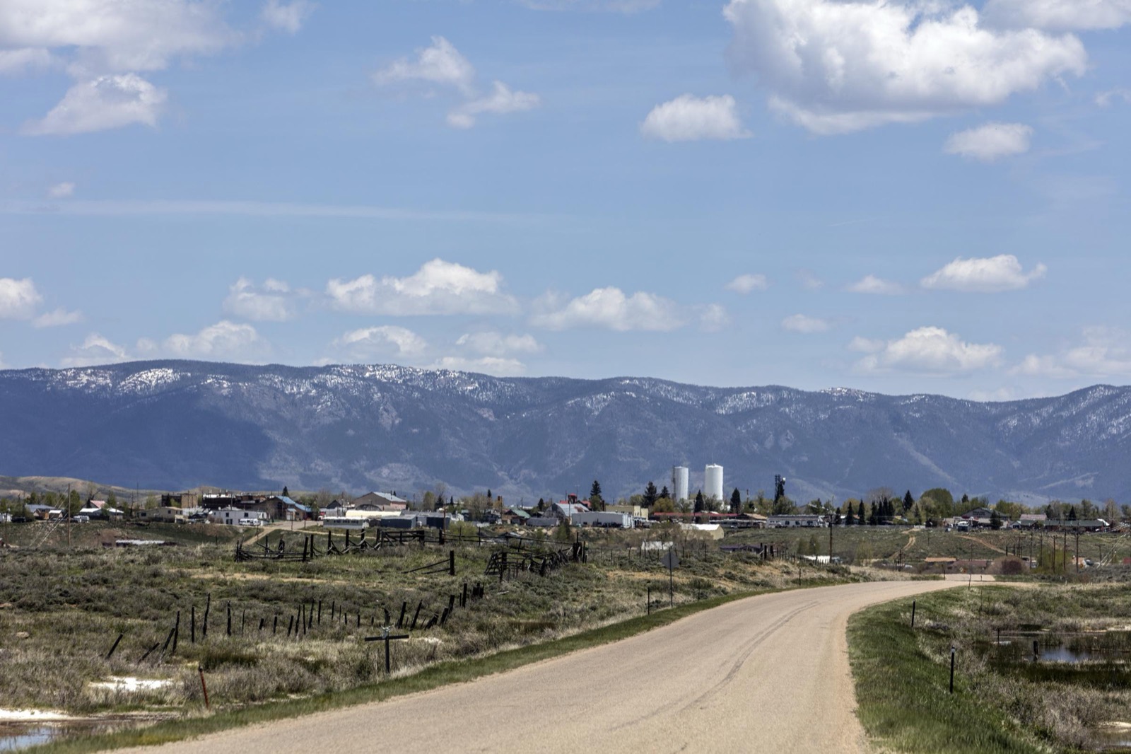 A dirt road curves through sagebrush toward the small ranching town of Walden, Colorado, with snow-dusted mountains behind it.